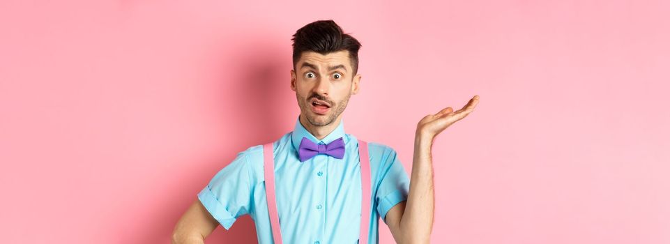 Confused guy cant understand what happening, looking with wtf face, raising hand and staring at camera shocked, standing over pink background.