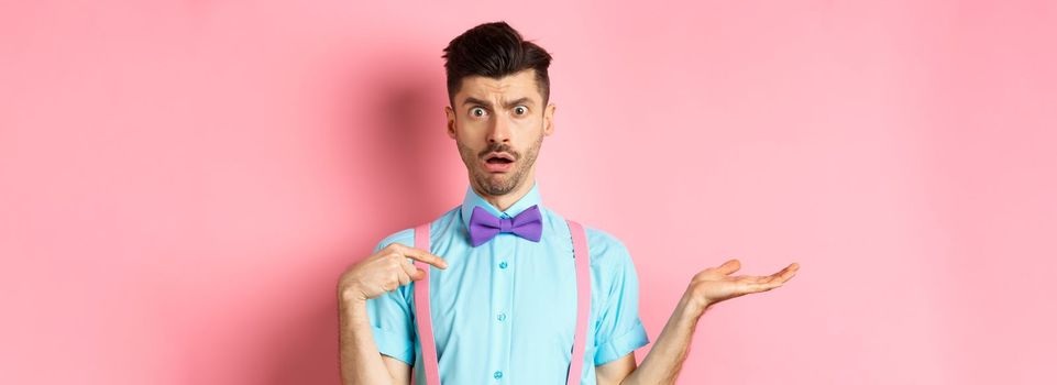 Shocked and confused guy pointing at himself with disbelief, being accused, standing on pink background in bow-tie and suspenders.