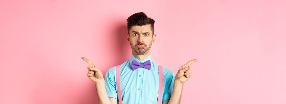 Indecisive young man pucker lips and frowning, pointing fingers sideways at two things, cant decide, standing puzzled over pink background.