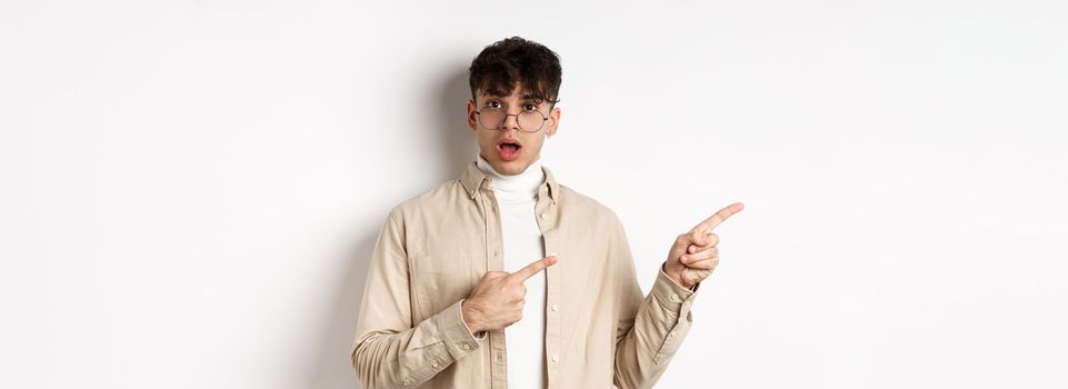 Portrait of young man in glasses drop jaw impressed, pointing fingers left at empty space with amazed face, standing on white background.