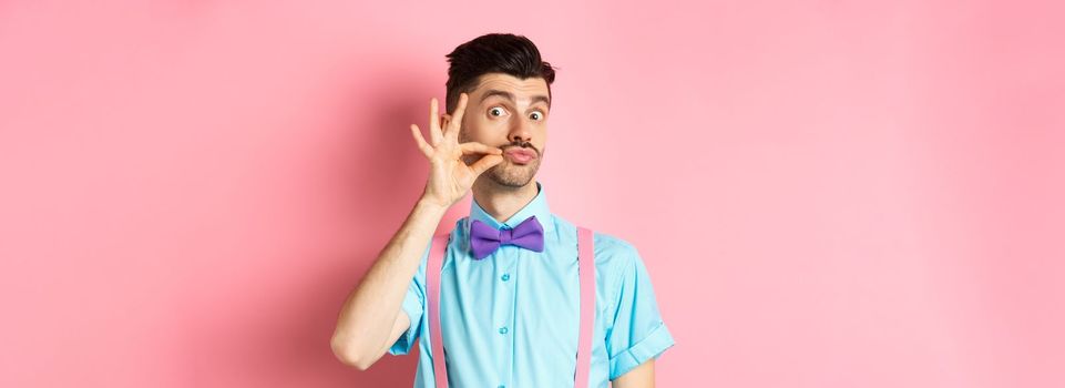 Funny young man touching his french moustache and pucker lips, looking silly at camera, standing in bow-tie and suspenders on pink background.