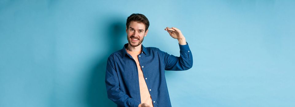 Handsome caucasian guy showing something big, shaping large size object with hands and smiling, standing on blue background.
