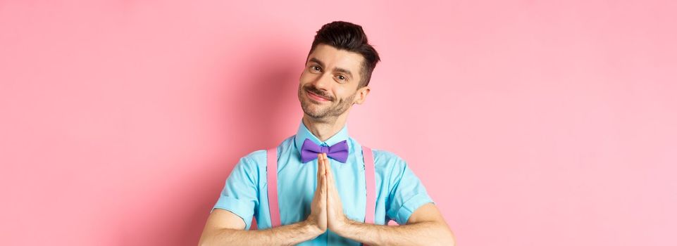 Smiling young guy in bow-tie say thank you, looking with gratitude and holding hands in namaste gesture, standing over pink background. Copy space