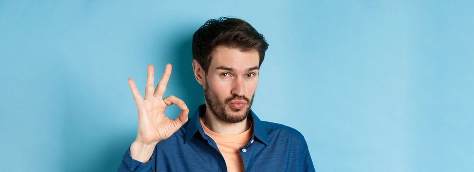 Image of silly young man pucker lips and show okay gesture, praise good thing, compliment excellent choice, standing on blue background.