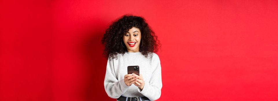 Image of young woman with curly hair, reading message on smartphone and smiling happy, receiving good news online, standing on red background.