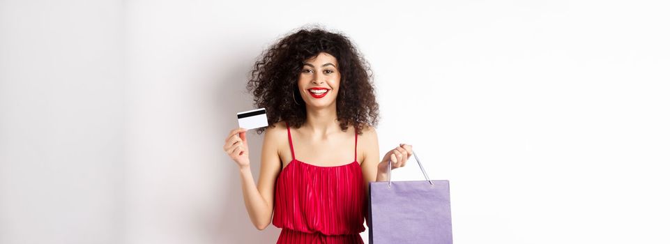 Beautiful female model with curly hair, red dress, showing shopping bag and plastic credit card, white background.