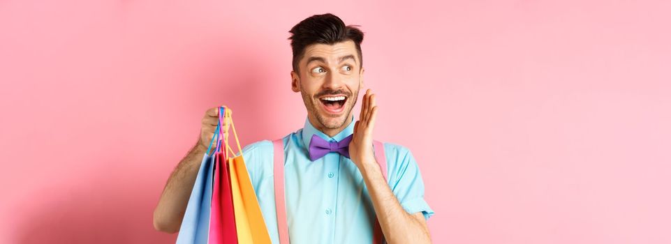 Surprised happy man screaming of joy, looking aside at logo and holding shopping bags, standing over pink background.