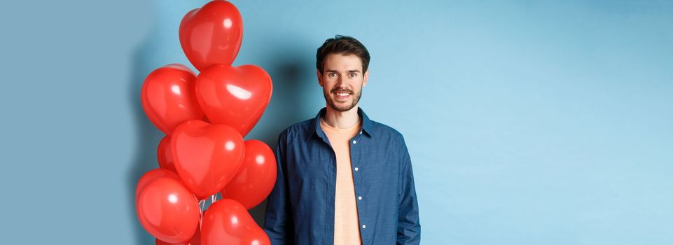 Cute boyfriend looking happy and smiling, standing near Valentines day heart balloons on blue background.