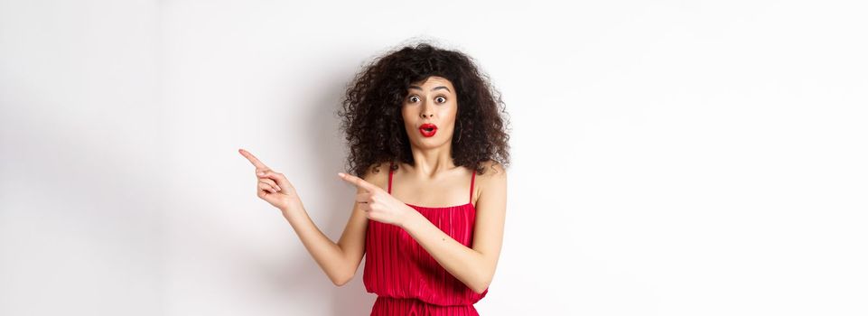 Surprised woman in red dress and makeup pointing fingers right, showing logo and look intrigued, standing on white background.