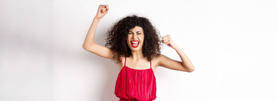 Cheerful emotive woman with curly hair, red dress, raising hands up and chanting, rooting for team, shouting wanting to win, standing on white background.