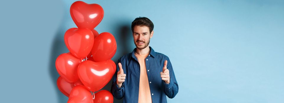 Happy valentines day. Confident man pointing fingers at camera and smiling, standing with balloons on blue background.