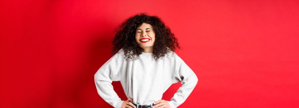 Excited beautiful woman laughing and smiling carefree, enjoying life, standing in sweatshirt against red background.