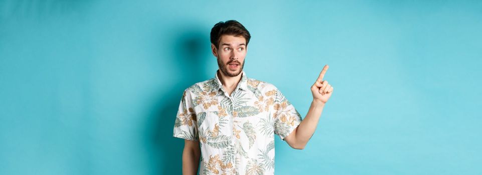 Intrigued man in hawaiian shirt, looking and pointing left at something interesting, blue background. Concept of tourism and vacation.