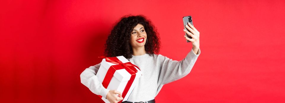 Happy young woman taking selfie with her valentines day gift, holding present and photographing on smartphone, posing on red background.