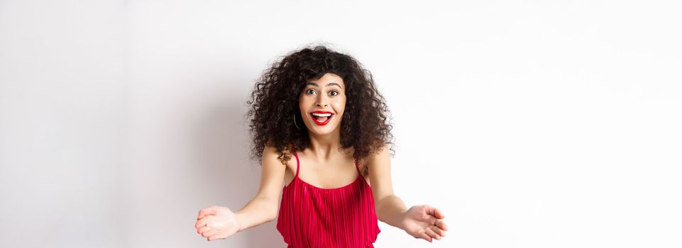 Portrait of happy caucasian woman in red dress and makeup, stretch out hands to beckon someone, inviting for hug, receiving surprise gift, standing on white background.