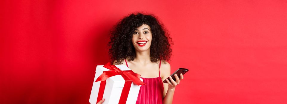 Online shopping and Valentines day. Beautiful young woman holding smartphone and lovers gift, looking surprised and happy at camera, red background.