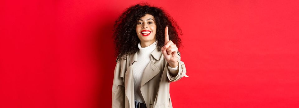 Cheerful woman in trench coat, showing number one finger and smiling, standing on red background.
