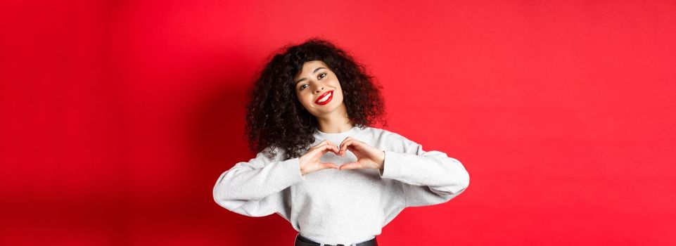 Beautiful young woman with curly hair showing heart gesture, say I love you and smile romantic at camera, standing on red background.