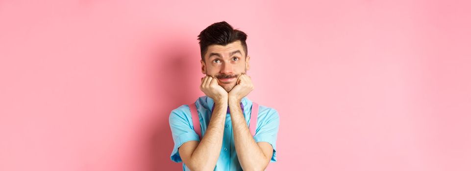 Silly young man leaning on hands and looking up, dreaming of something, imaging things on pink background.