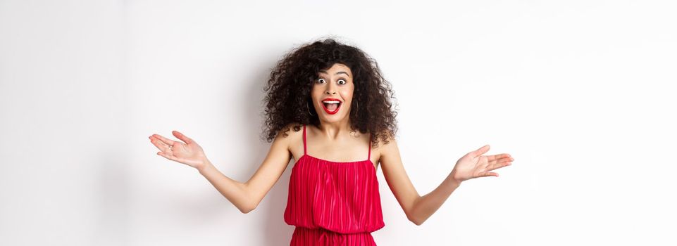 Excited young woman in red dress, spread hands sideways and screaming surprised, standing happy on white background.