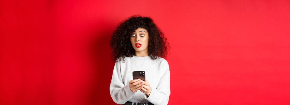 Shocked woman stare at smartphone screen with popped eyes, reading strange message, standing on red background.