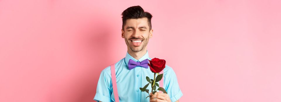 Happy man showing tongue and smiling, holding red rose for girlfriend on Valentines day, enjoying romantic date, pink background.