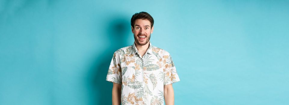Summer holiday. Handsome happy man in hawaiian shirt looking amused, smiling at camera, standing on blue background.
