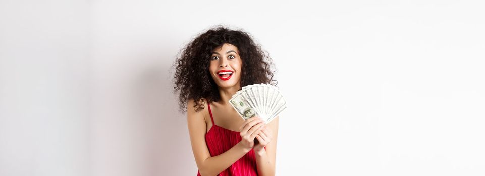 Excited woman in red dress winning money, showing dollar bills and smiling happy, standing on white background.
