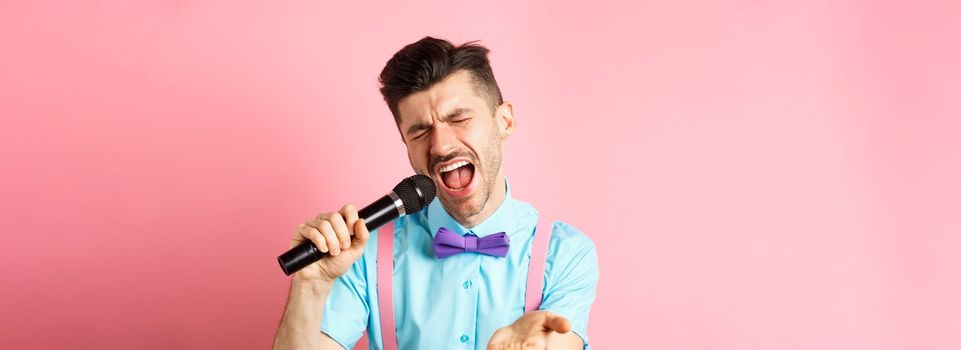 Party and festive events concept. Passionate singer holding microphone and pointing hand at you, singing about love, standing on pink background.