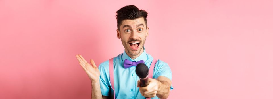 Cheerful young man, show host in bow-tie giving you microphone and smiling, entertain people on festive celebration events, performing at holidays, standing on pink background.