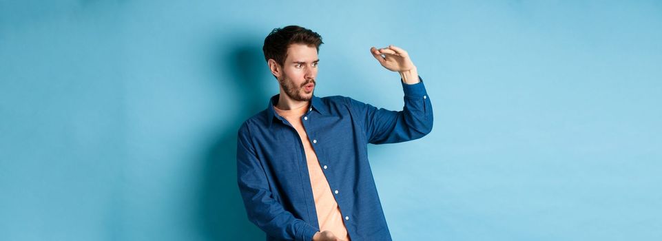Impressed caucasian guy staring at empty space with big object, shaping large size, standing on blue background.