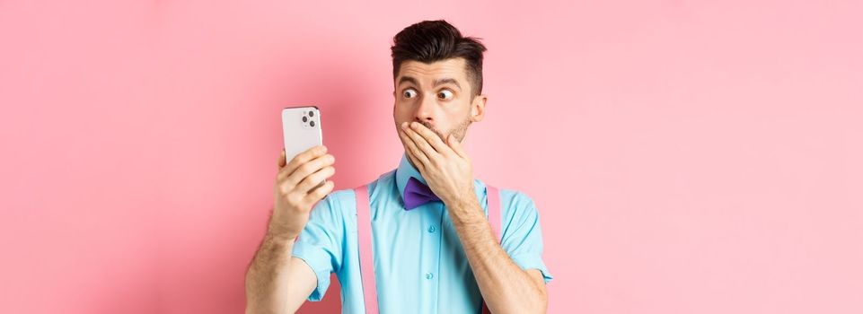 Technology concept. Silly young man looking shocked at smartphone screen while having video chat, standing on pink background and cover mouth with hand.