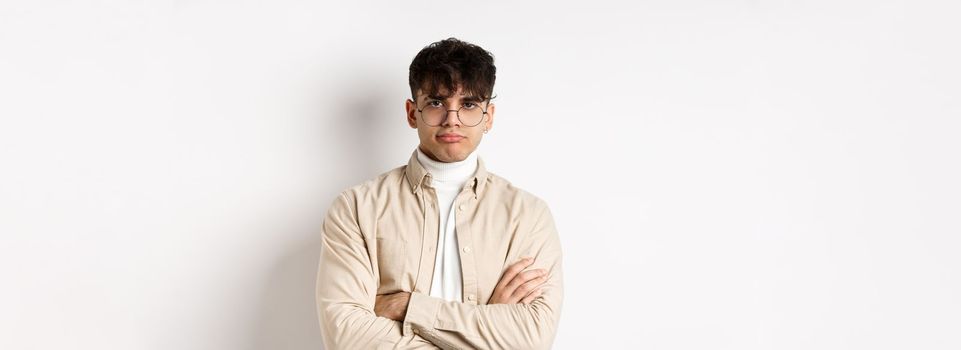 Image of stylish hipster guy in glasses feeling bored, looking unamused with pokerface, cross arms on chest and stand reluctant against white background.