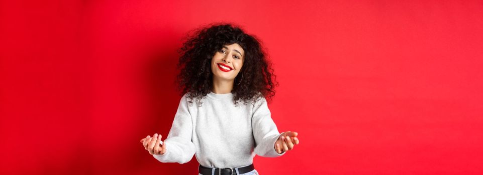 Pretty woman with curly haircut, extending hands and smiling with love and tenderness, inviting to come closer, reaching arms for hug, taking something into her arms, red background.