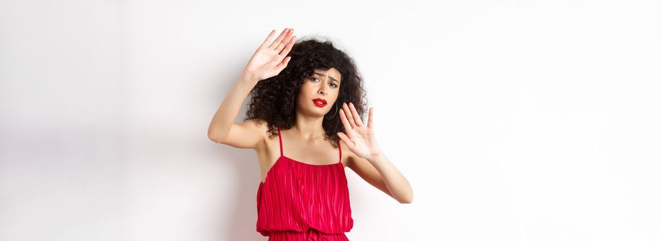 Young woman with curly hair and red dress, asking to stop, block someone, raising hands defensive, protecting herself, standing against white background.