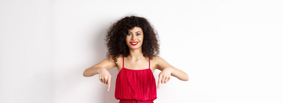 Attractive female model in red dress and makeup, pointing fingers down and smiling, showing advertisement, standing over white background.