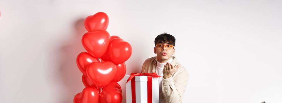 Valentines day and romance concept. Romantic modern man holding special gift for lover and sending air kiss at camera, standing near hearts balloons, white background.