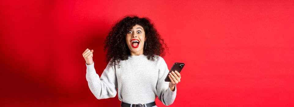 Excited caucasian woman with curly hair, chanting, winning online prize, holding smartphone and triumphing, standing on red background.