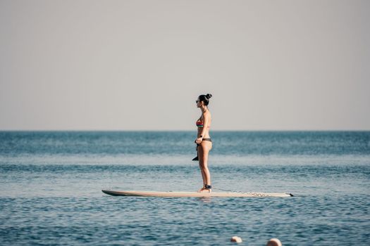 Sea woman sup. Silhouette of happy middle aged woman in rainbow bikini, surfing on SUP board, confident paddling through water surface. Idyllic sunset. Active lifestyle at sea or river