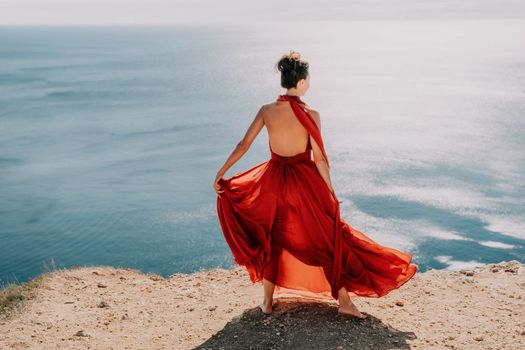 Side view a Young beautiful sensual woman in a red long dress posing on a rock high above the sea during sunrise. Girl on the nature on blue sky background. Fashion photo.