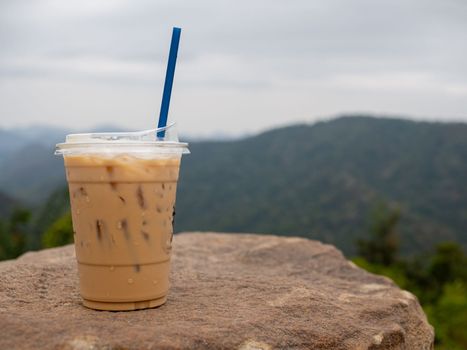 A glass of iced coffee is placed on a rock against a background of mountains and sky.