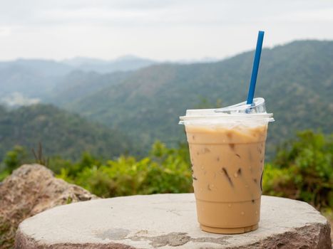 A glass of iced coffee is placed on a rock against a background of mountains and sky.