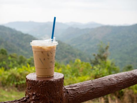 A glass of iced coffee is placed on the fence against a backdrop of mountains and sky.