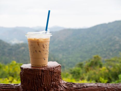 A glass of iced coffee is placed on the fence against a backdrop of mountains and sky.