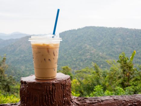 A glass of iced coffee is placed on the fence against a backdrop of mountains and sky.