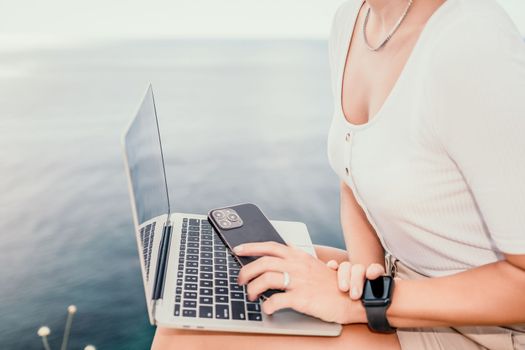 Digital nomad, Business woman working on laptop by the sea. Pretty lady typing on computer by the sea at sunset, makes a business transaction online from a distance. Freelance remote work on vacation