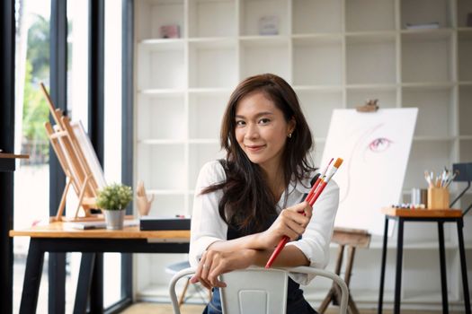 Smiling woman artist holding painting brush and sitting in her art studio.