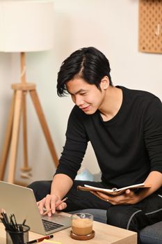 Young man holding book and using laptop computer in living room.