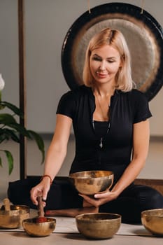 A woman plays a Tibetan singing bowl while sitting on a yoga mat against the background of a gong.