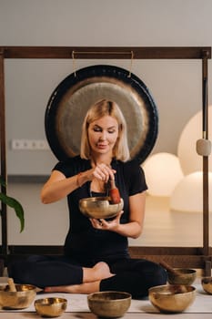 A woman plays a Tibetan singing bowl while sitting on a yoga mat against the background of a gong.